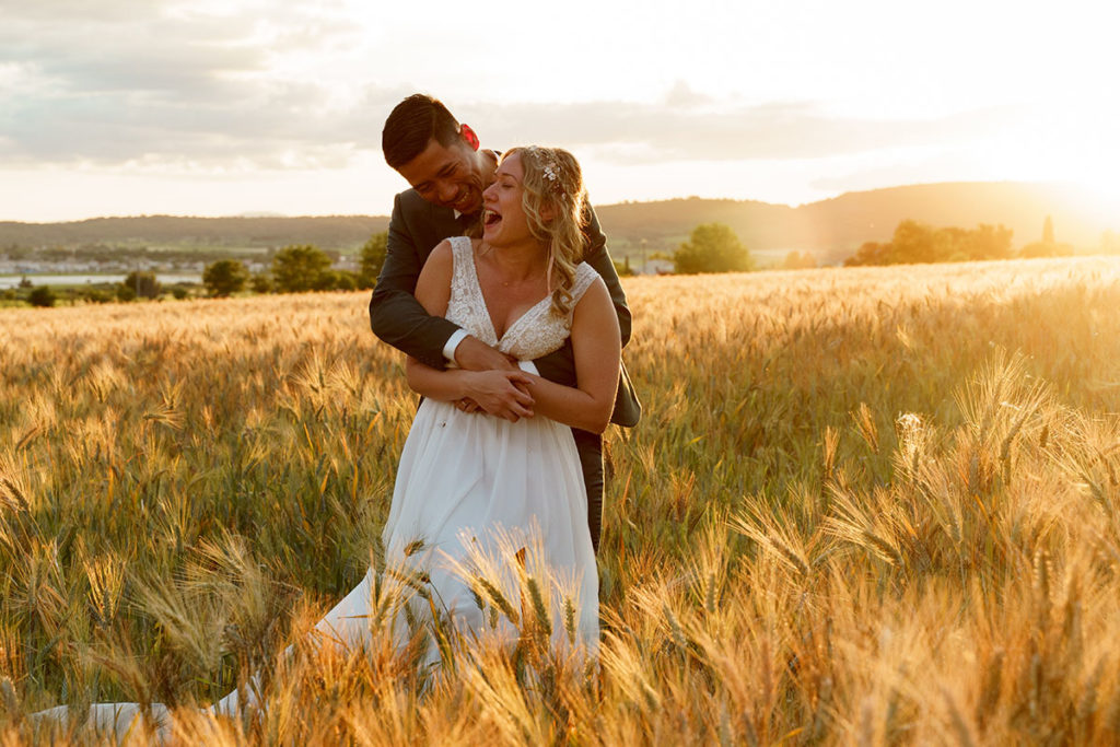 Photo de couple dans un champ de blé au soleil couchant, la mariée éclate de rire quand son chéri lui fait des chatouilles.